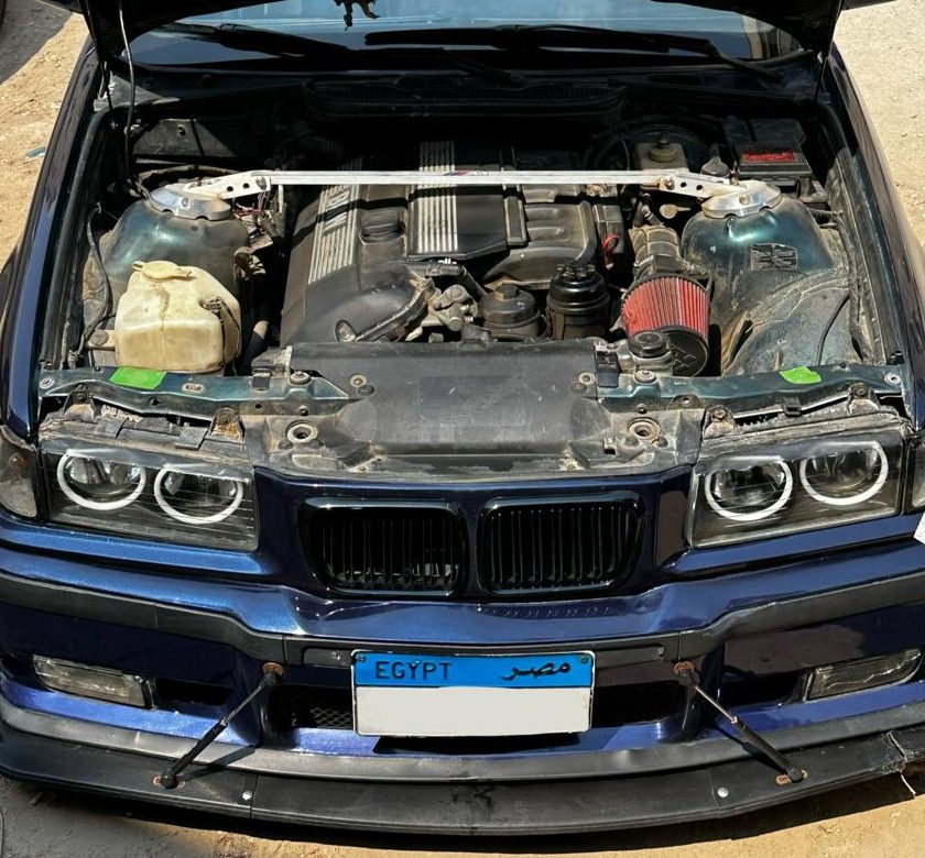 The E36 engine bay with the bonnet raised, showing a polished strut brace across the suspension towers, a red cone intake filter, and the four angel eye rings in the opened headlight housings below