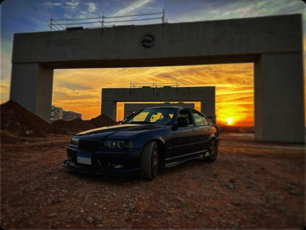 The blue E36 parked on gravel beneath a concrete overpass at sunset, silhouetted against an orange sky