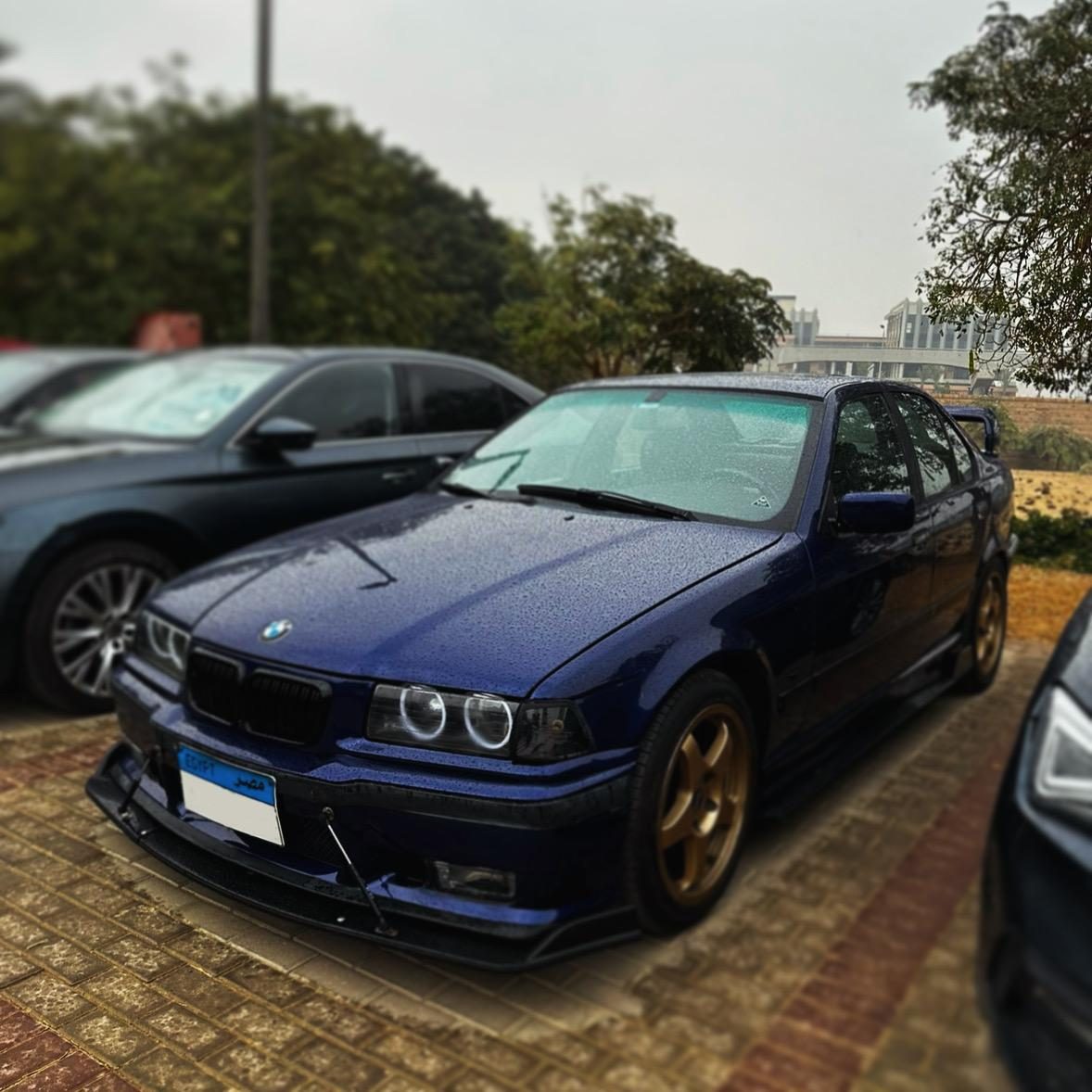 Metallic blue BMW E36 saloon parked on brick paving, seen from the front three-quarter, with gold split-spoke wheels, angel eye headlight rings and a black front splitter, beaded with rain